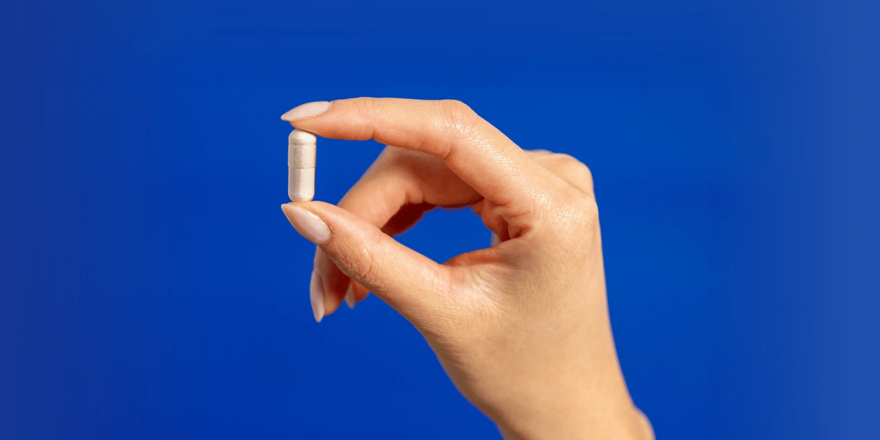 Primo piano di una mano che tiene una capsula beige di integratore alimentare tra il pollice e l’indice su uno sfondo blu.